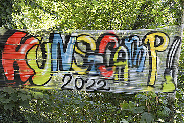 Auf diesem Bild ist ein Schriftzug der mit Graffiti auf weißer Folie aufgetragen ist. Dort ist "Kunstcamp 2022" aufgesprüht.