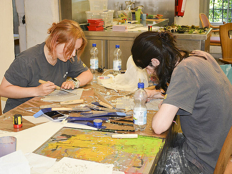 Auf dem Bild sind zwei Teilnehmerinnen des Kunstcamps zu sehen. Beide sitzen an einem Tisch auf dem sie mit Stanzwerkzeugen Vorlagen zum Linoleumdruck erstellen. 