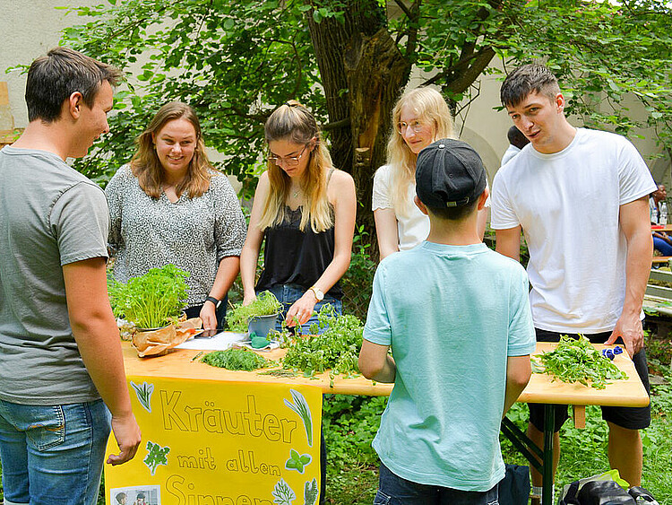 Das Foto zeigt einen weiteren Stand des Schulgartenfest. Ingesamt stehen sechs Personen um den Tisch. Auf dem Tisch sind verschiedene Kräuter verteilt. An dem Tisch ist ein Plakat mit der Aufschrift "Kräuter mit allen Sinnen" besfestigt.