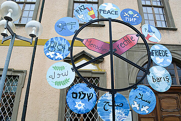 Auf diesem Bild ist eine Kunstinstallation zur Tagung zu sehen. Diese besteht aus einem Metallrahmen als Friedenssymbol, dort angebracht sind kleine Tafeln mit dem Wort "Friede" in verschiedenen Sprachen.