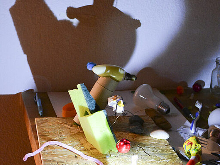 Das Bild zeigt ein Schattenspiel das als Kunstopbjekt im Kunstcamp erstellt wurde. Dafür wurden veschiedene Materialien, wie Schwämme, Draht und andere "Schrott"-Objekte auf einer Holzplatte befestigt und über eine Lampe an die Wand projeziert. 