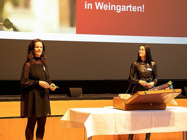 Zwei Frauen der Studienberatung stehen hinter einem Tisch mit einem Koffer und halten einen Vortrag.